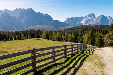 Scenic hiking trail along wooden fence on alpine meadow on Nemes Alm (Rifugio Malga Nemes) in Carnic Alps, South Tyrol, Italy. View of massive mountain ridges of majestic untamed Sexten Dolomites - obrazy, fototapety, plakaty