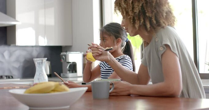 Biracial mother and daughter eating breakfast cereal in sunny kitchen, copy space, slow motion