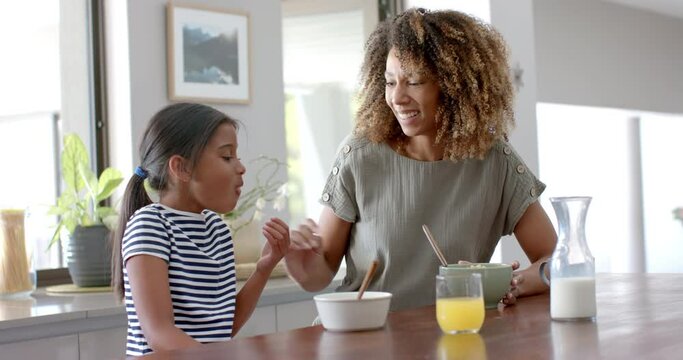 Happy biracial mother and daughter eating breakfast cereal in sunny kitchen, slow motion