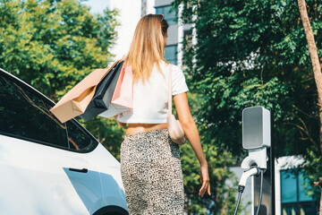 Young woman travel with EV electric car charging in green sustainable city outdoor garden in...