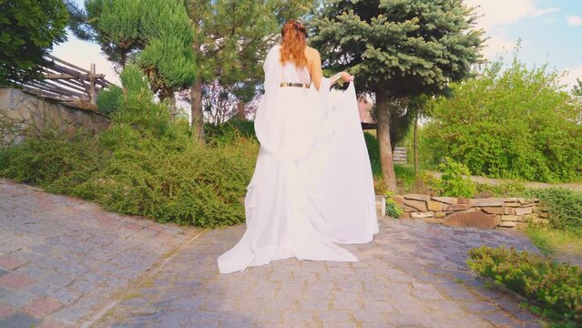 Bride woman walking along the path back rear view in spring garden fresh plants, green tree summer nature day sun light. Sexy girl queen antique greek style white silk long dress. Fantasy woman art 4k