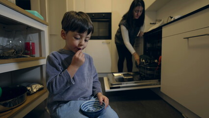 Family Everyday Life - Child Snacking on Blueberries While Mom Tidies Kitchen. young boy wearing pajamas in the evening