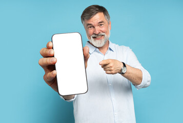 Handsome Excited Mature Man Showing Pointing At Empty Smartphone Screen Posing Over Light Blue Studio Background, Smiling To Camera. Check This Out, Cellphone Display Mock Up