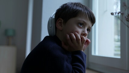 Thoughtful small boy with hand in chin daydreaming at home by window. Pensive child thinking deeply absorbed in introspective thoughts