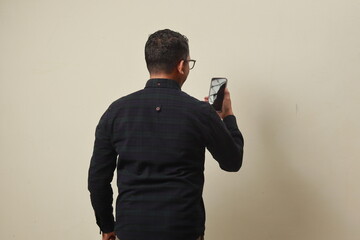 Rear view of adult man wearing long-sleeved shirt looking at mobile phone isolated on black. Space...