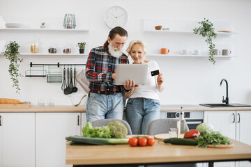 Old Caucasian couple using laptop and credit card to buy food products to prepare breakfast in modern spacious kitchen background. - obrazy, fototapety, plakaty