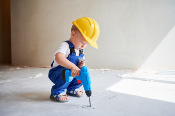 Child construction worker drilling floor with toy electric drill while crouching down near the wall in apartment. Kid in safety helmet working with power drilling tool at home during renovation. - obrazy, fototapety, plakaty
