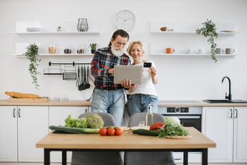 Old Caucasian couple using laptop and credit card to buy food products to prepare breakfast in modern spacious kitchen background.