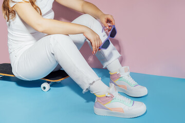 No face female in white outfit and retro style high-top multicolor sport sneakers shoes with sunglasses sitting on skateboard on blue pink background. Vintage retro fashion style of 80s - 90s vibes - obrazy, fototapety, plakaty