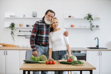 Happy old couple husband and wife in casual clothes hugging while preparing healthy salad in modern kitchen. Gray-haired man hugs an old woman on a weekend morning.