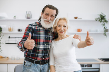 Portrait of happy Caucasian pensioners gray-bearded husband and wife hugging on light kitchen...