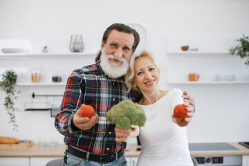 Respectful positive senior couple gray-bearded man and blonde woman having fun with vegetables: broccoli and tomatoes, preparing healthy vegetarian salad in the kitchen.