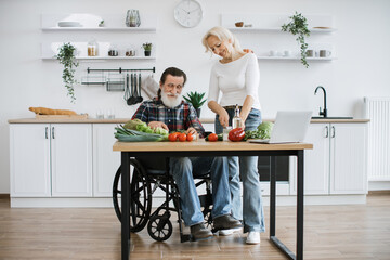 Happy old married man in wheelchair and wife prepare healthy salad, cutting thin slices of greenery vegetables and tomatoes with knife against background of spacious kitchen.