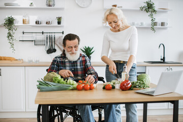 Happy old married man in wheelchair and wife prepare healthy salad, cutting thin slices of greenery vegetables and tomatoes with knife against background of spacious kitchen.