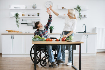 Old married man in wheelchair and wife prepare healthy salad, cutting thin slices of greenery vegetables and tomatoes with knife against background of spacious kitchen giving each other high five.