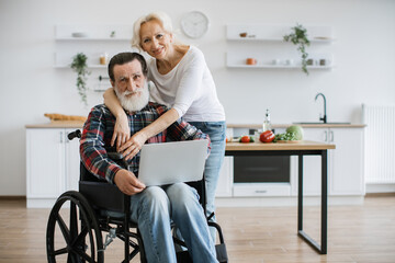 Charming old couple of man in wheelchair with laptop and wife hugging him on background of bright modern kitchen, looking at camera.