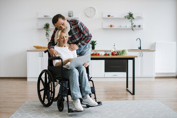 Full length portrait of charming old couple of woman in wheelchair with laptop and husband hugging...