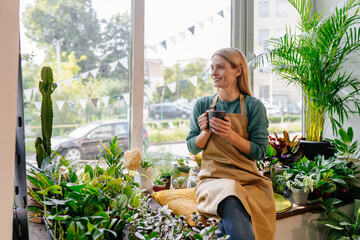 Business female owner relaxing drinking coffee at flower shop sitting surrounded by plants at...