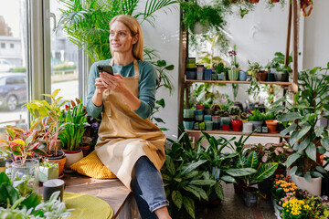 Beautiful smiling blonde middle age woman sitting with phone near window among flowers and browsing messages or websites in cell phone in interior of store with plants. - Powered by Adobe