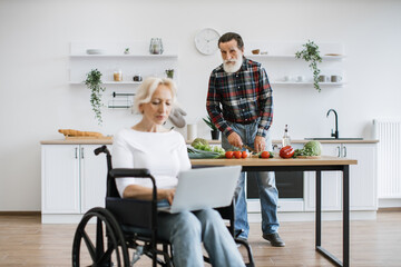 Old caucasian husband with gray beard prepares healthy salad in kitchen while his wife woman sitting in wheelchair and working on laptop.