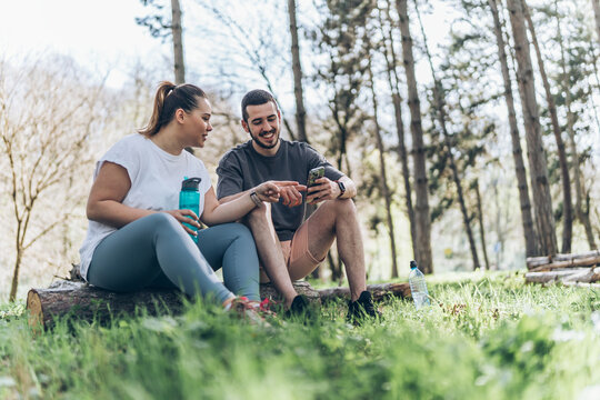 Sitting on a fallen tree trunk, the couple, including the determined overweight female, catch their breath, their laughter blending with the rustling leaves as they enjoy a post-workout moment.