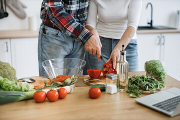 Cropped view of hands of positive retired couple cutting pepper while holding knife together in hands and putting pieces into glass bowl.