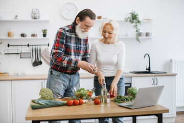 Elderly man with gray beard tears lettuce and herbs into glass bowl while wife cutting peppers in small pieces. Old retired couple preparing fresh healthy salad in modern kitchen.