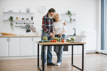 Caucasian old couple spends free time cooking breakfast in modern light kitchen. Seniors husband and wife mix chopped fresh vegetables in glass bowl while prepare delicious healthy salad.