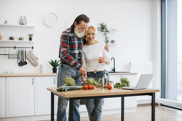 Caucasian old couple spends free time cooking breakfast in modern light kitchen. Seniors husband and wife mix chopped fresh vegetables in glass bowl while prepare delicious healthy salad.