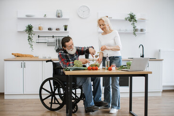 Old couple with disability spends free time cooking breakfast in modern light kitchen. Seniors husband in wheelchair and wife mixing chopped vegetables in bowl while prepare delicious healthy salad.