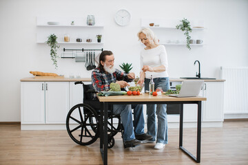 Seniors husband in wheelchair and wife mixing chopped vegetables in bowl while prepare delicious healthy salad. Old couple with disability spend free time cooking breakfast in modern light kitchen.