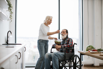 Happy elderly disabled couple having breakfast in modern kitchen with panoramic windows. Devoted woman feeds man in wheelchair with fresh healthy salad.