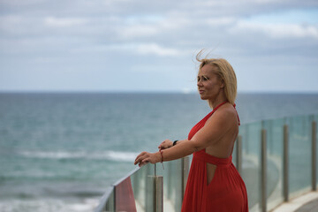 Mature, beautiful, blonde woman in a red dress, leaning on a glass railing looking out to sea while the wind stirs her hair. Concept beauty, fashion, trend, travel, maturity.