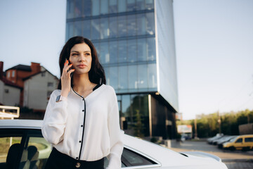Beautiful young happy caucasian business woman standing near her new car and using smartphone. Buying and renting a car. Travel, tourism, recreation.