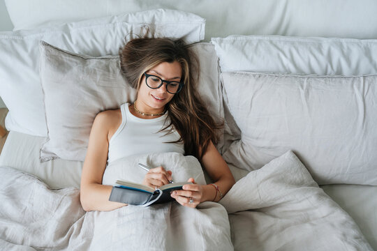 Relaxed young woman writing in a journal while lying in bed, enjoying a quiet moment in a peaceful bedroom