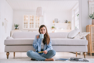 Cheerful woman sipping coffee on the floor, with a cozy living room in the background, radiating relaxation