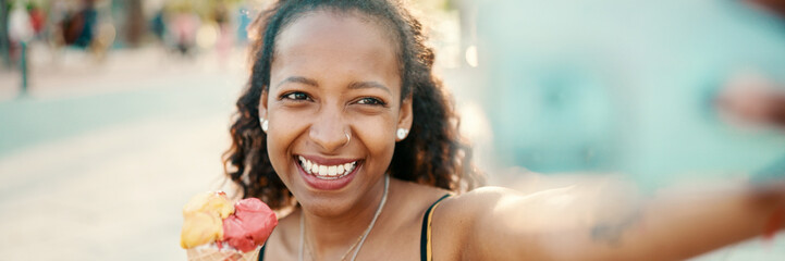 Closeup of smiling young woman with long curly hair with ice cream in her hands making a video call...