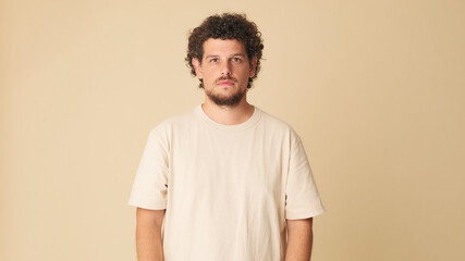 Amazed guy with curly hair dressed in beige t-shirt looking at the camera with big eyes expressing...