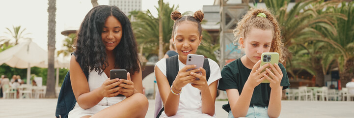 Three girls friends pre-teenage are sitting on the waterfront using mobile phone. Three teenagers...