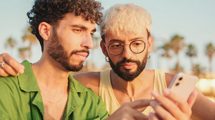 Close-up of young gay couple sitting hugging on the beach, smiling, talking and using mobile phone