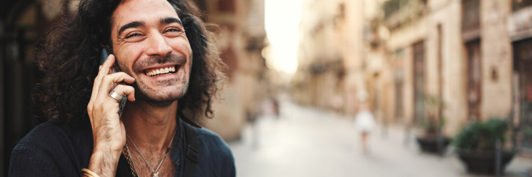 Fototapeta Young attractive italian guy with long curly hair and stubble is using mobile phone at old buildings background. Stylish man with an earring in his ear and lot of chains emotionally talking on phone