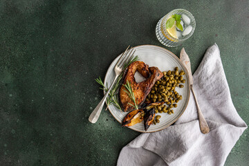 Sunlit healthy lunch setup featuring BBQ salmon steak seasoned with spices alongside roasted lemon and a serving of fermented capers