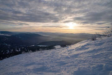 View from the top of the ski slope against the backdrop of mountains and forest under the evening sky.