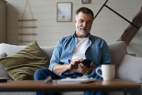 Happy smiling senior man using smartphone device while sitting on sofa at home