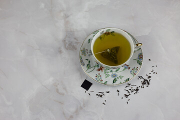 Obraz na płótnie Canvas Bag of green tea with water in a porcelain cup and saucer with a picture of color stands on a white marble table