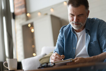 Senior man taking notes in notebook while sitting on the couch, gray-haired elderly man writing thoughts in notebook - obrazy, fototapety, plakaty