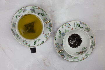 Fractional cups and saucers with a tea bag of green tea and black tea leaves on a white table. Choice of hot drink, black or green tea