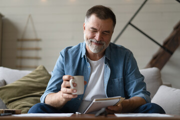 Senior man taking notes in notebook while sitting on the couch, gray-haired elderly man writing...