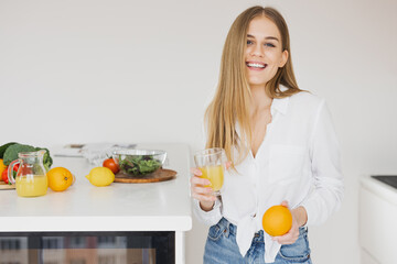 A happy cute blonde woman drinks orange juice in the kitchen - obrazy, fototapety, plakaty