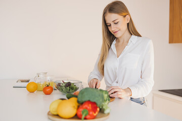 A happy cute blonde woman is preparing a salad in the kitchen